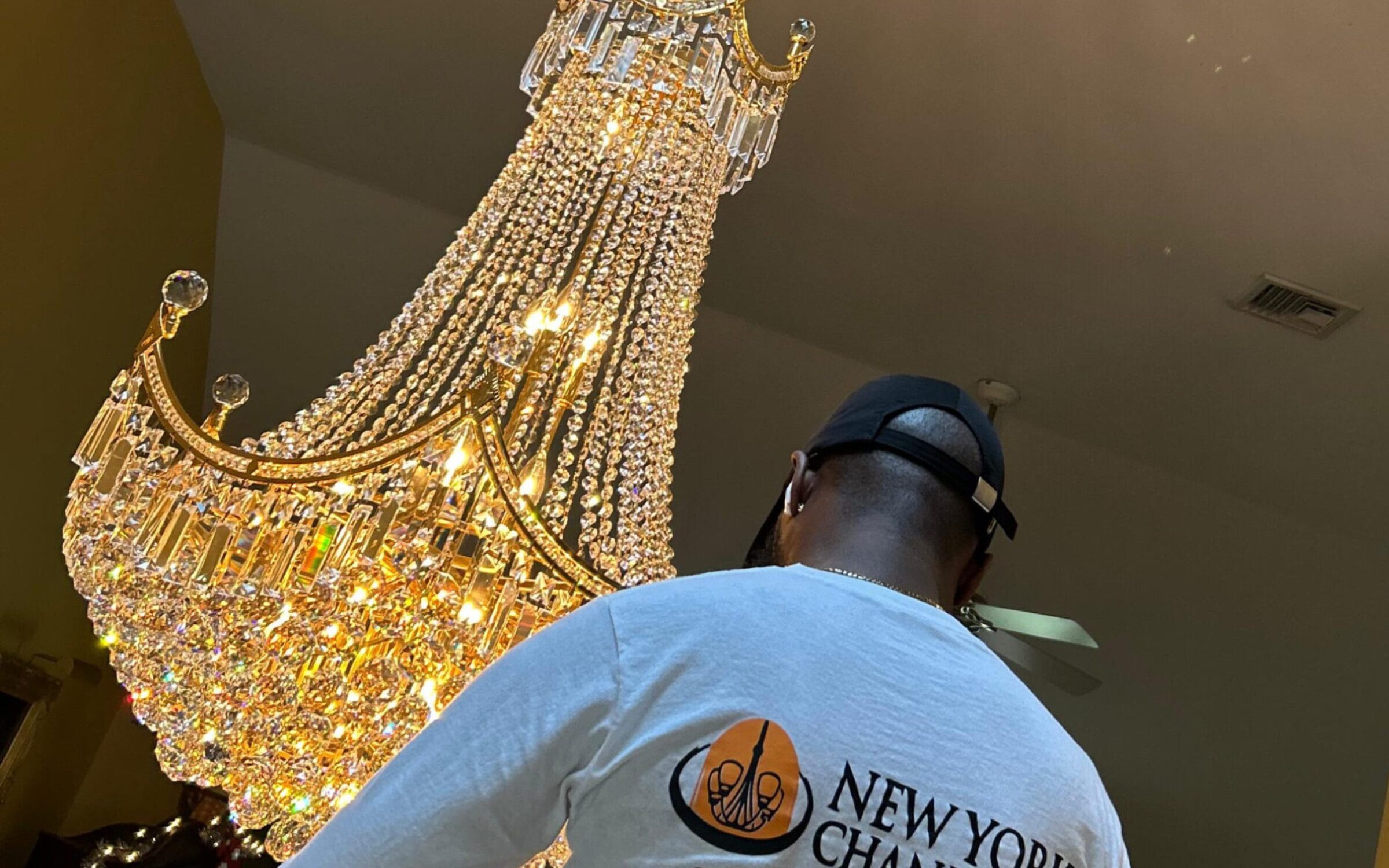 Person standing under a crystal chandelier wearing a shirt with "New York Chandelier" and phone number