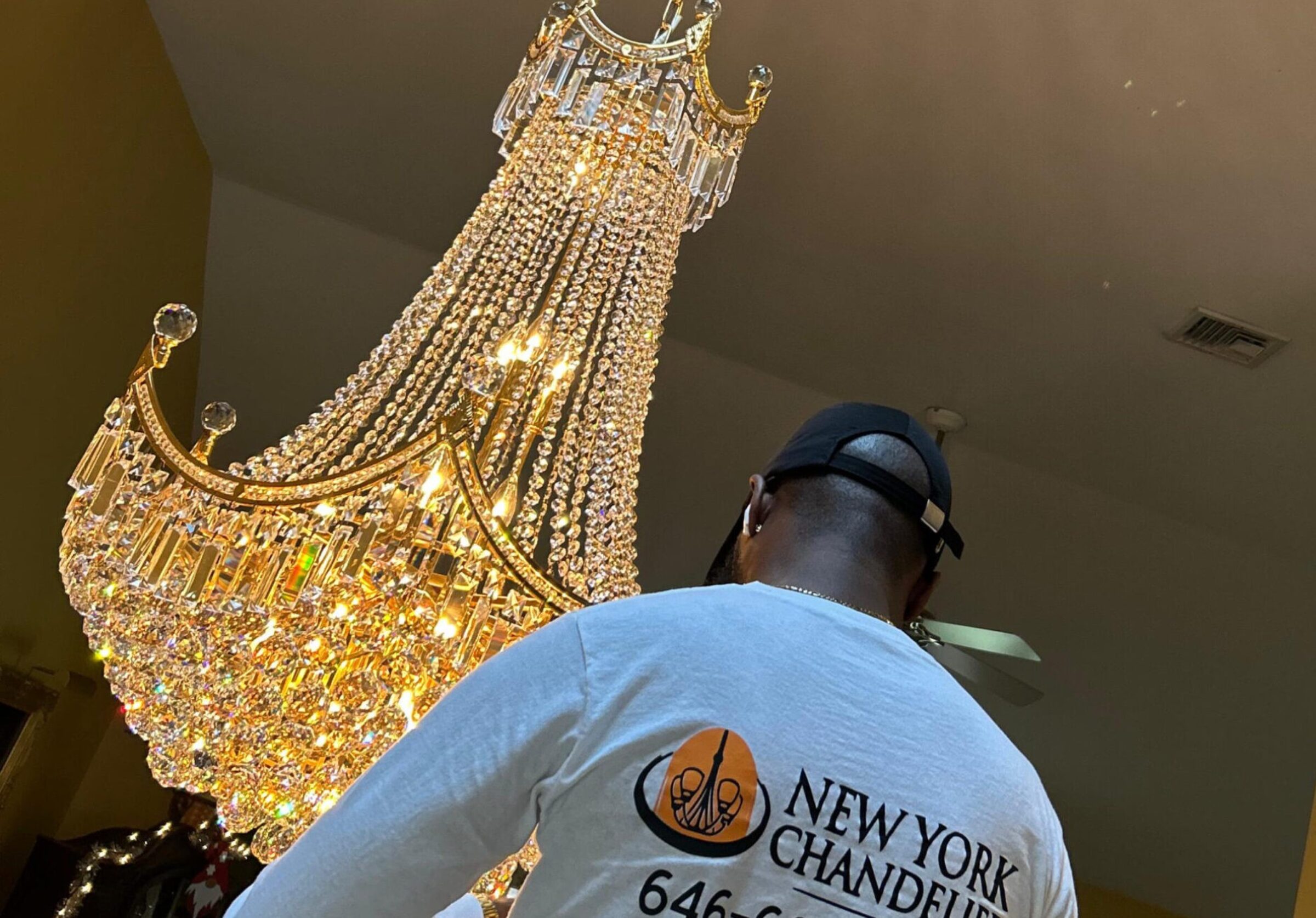 Person standing under a crystal chandelier wearing a shirt with "New York Chandelier" and phone number